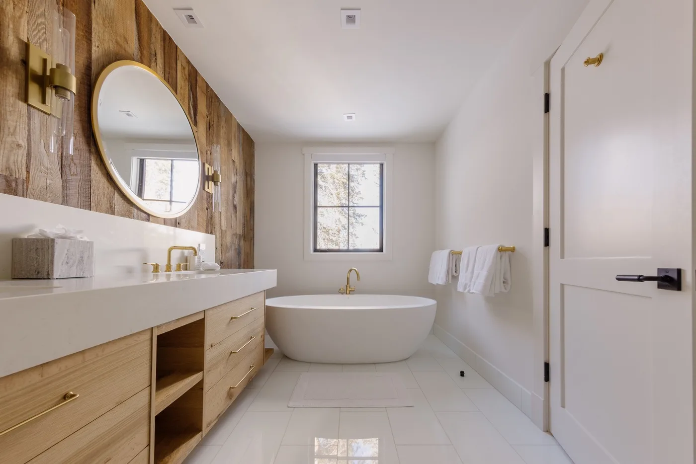 Un baño luminoso cuenta con una bañera exenta bajo una ventana y un tocador largo.
