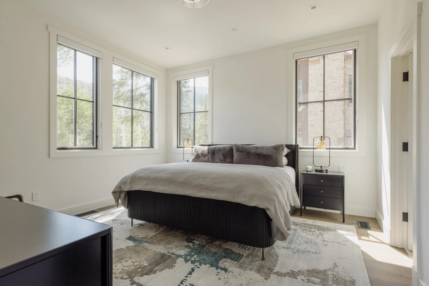 Un dormitorio luminoso con una cama, una mesita de noche y ventanas grandes.