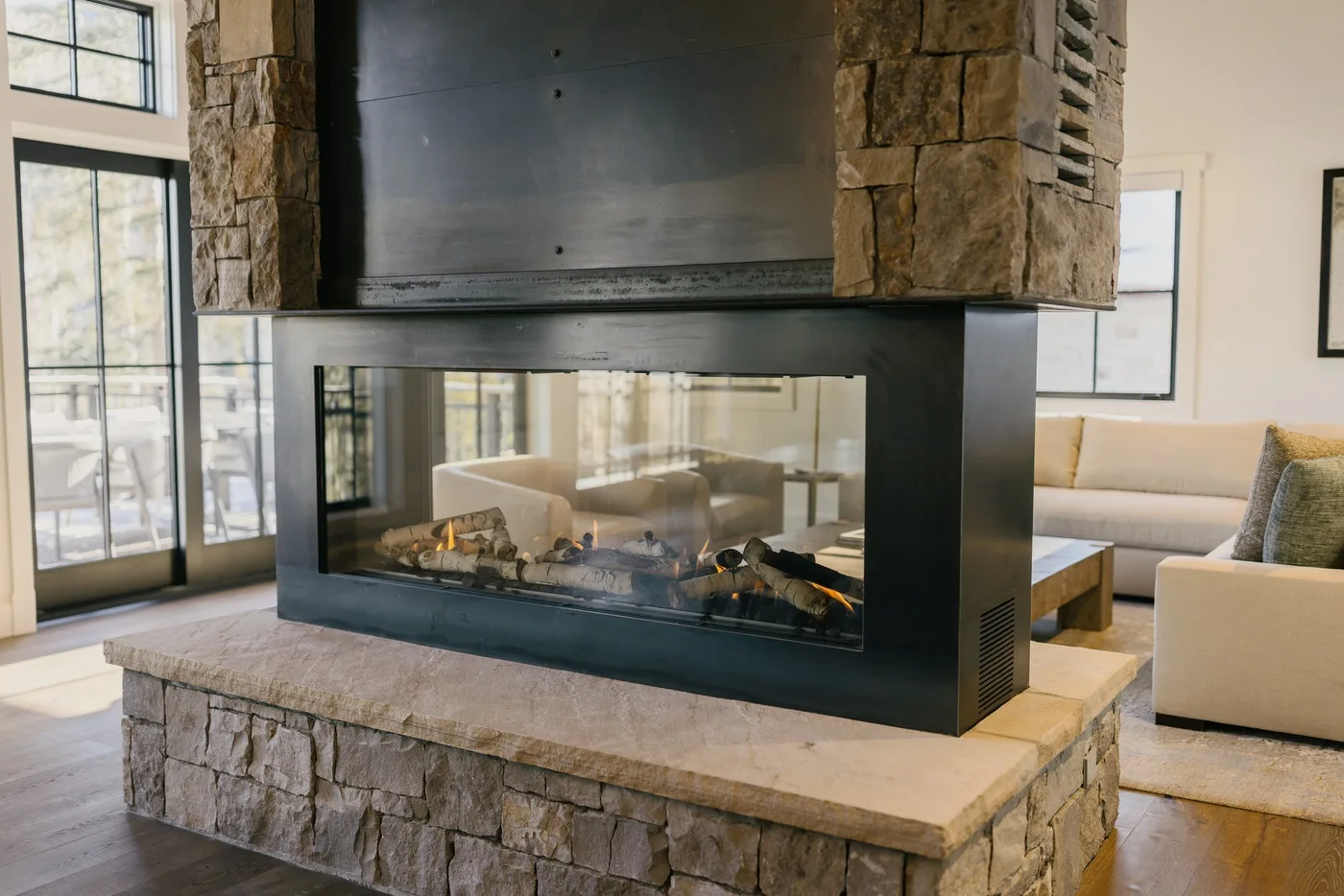 Una chimenea en una sala de estar está entre ventanas altas y asientos de tonos claros.