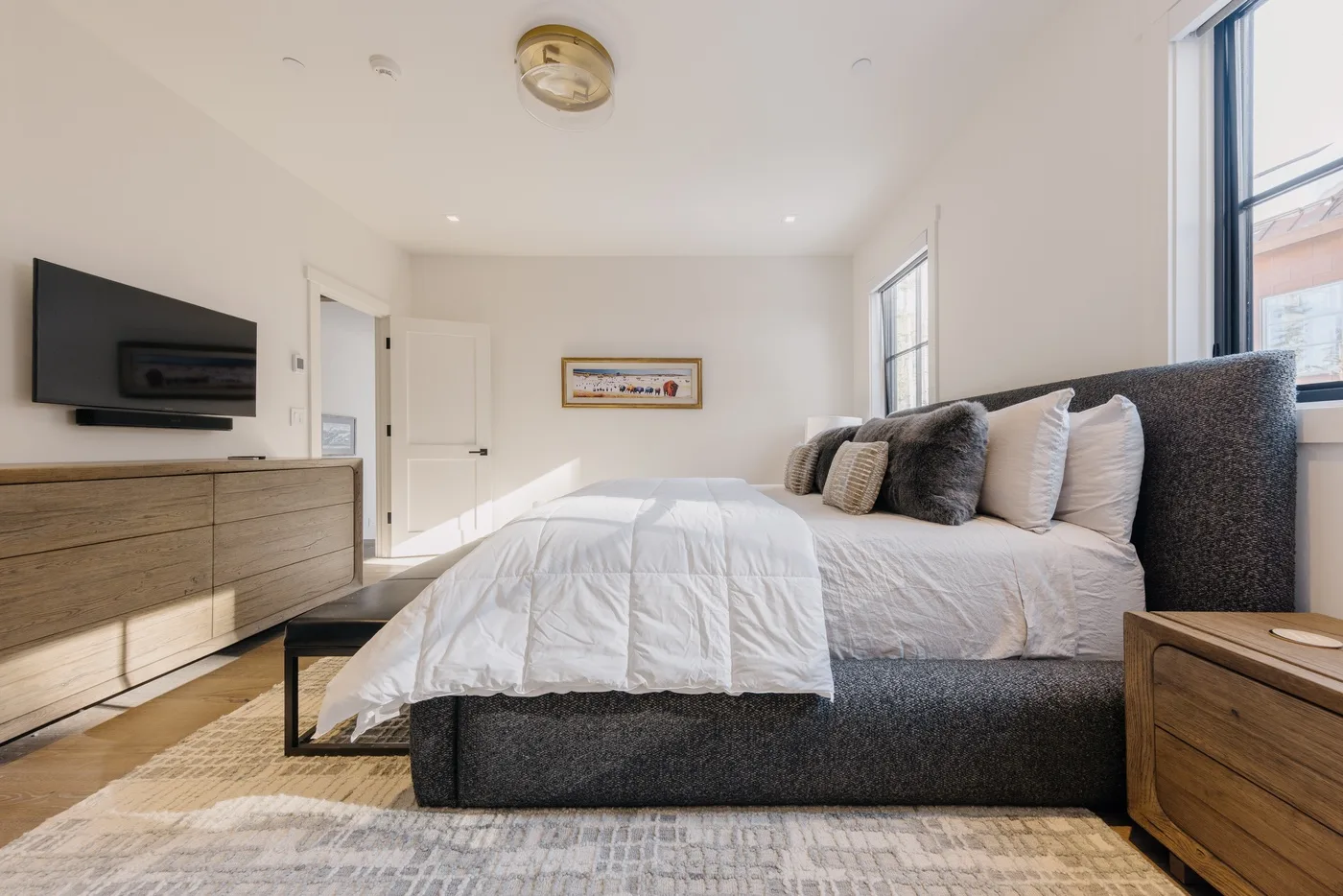 Un dormitorio luminoso con una cama tendida, un televisor en la pared y ventanas grandes.