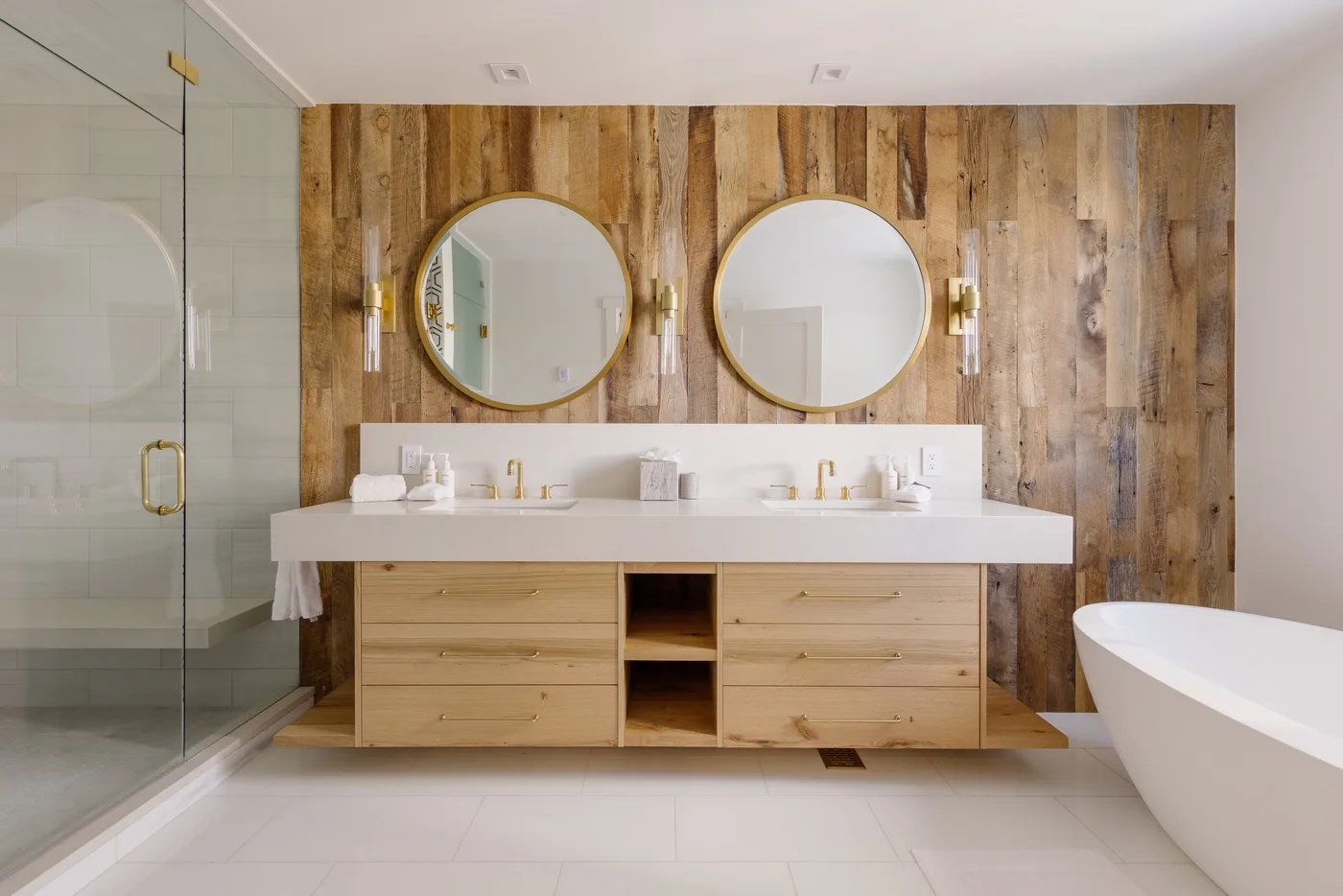 Un baño luminoso cuenta con un lavabo doble, espejos redondos y una bañera exenta.