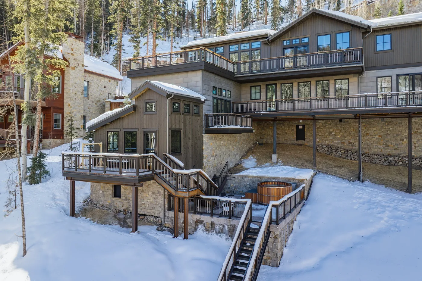 Un alojamiento de montaña cubierto de nieve, con terrazas de madera y una bañera de hidromasaje exterior.