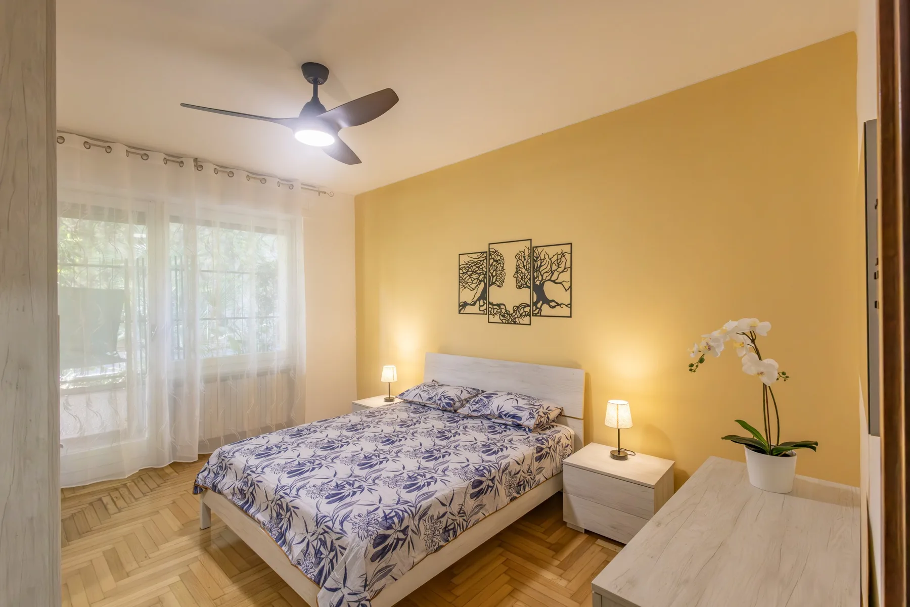 A bedroom with a double bed, patterned bedding, and a yellow accent wall.