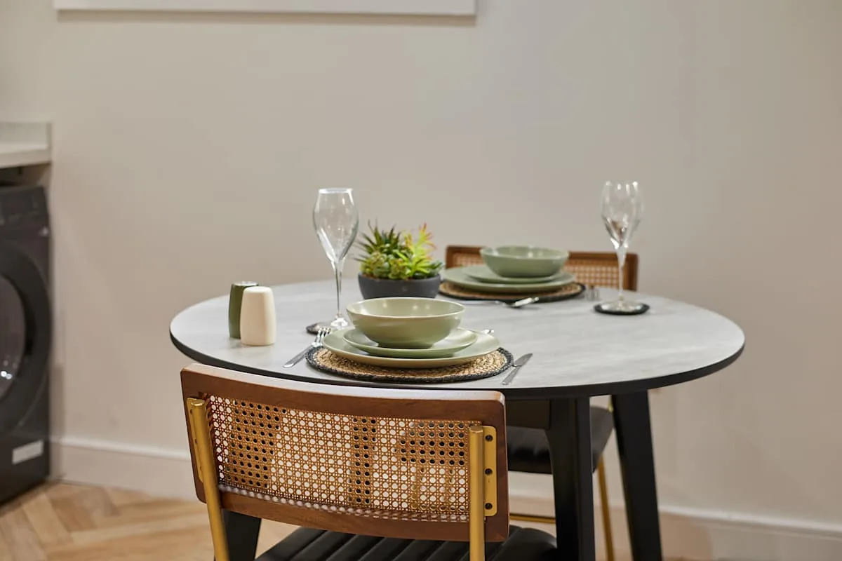 Una mesa redonda está preparada con platos y copas junto a una silla con respaldo tejido.