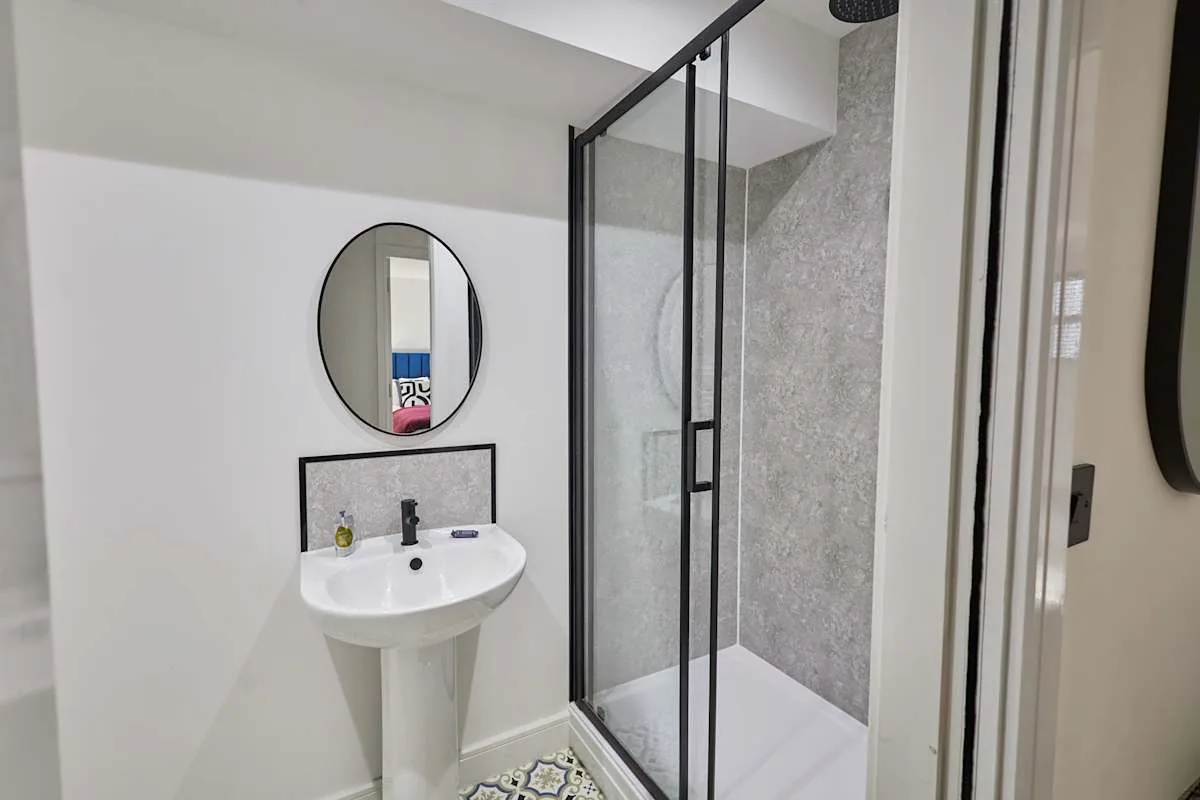 Un baño con lavabo de pedestal, espejo redondo y ducha de vidrio.