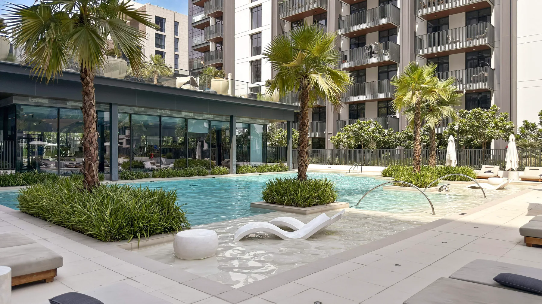 Una piscina al aire libre está rodeada de palmeras y edificios de apartamentos modernos.