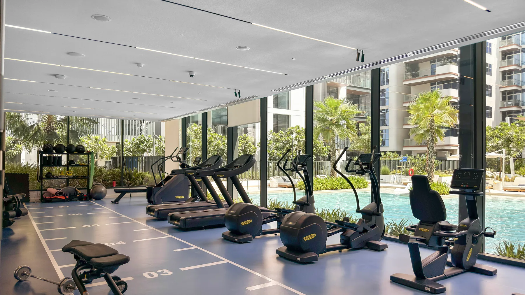 Un gimnasio luminoso con máquinas de ejercicio junto a grandes ventanales con vistas a una piscina.