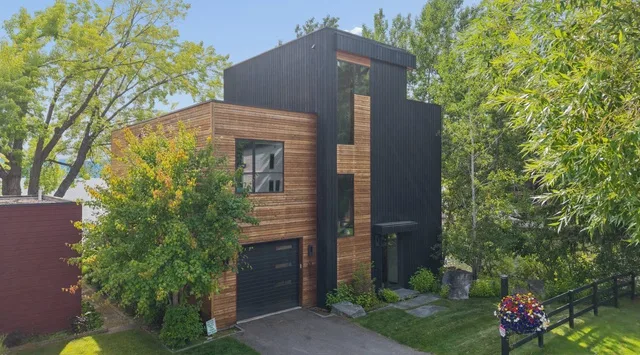 Una casa moderna con revestimiento oscuro y detalles de madera está rodeada de árboles frondosos.