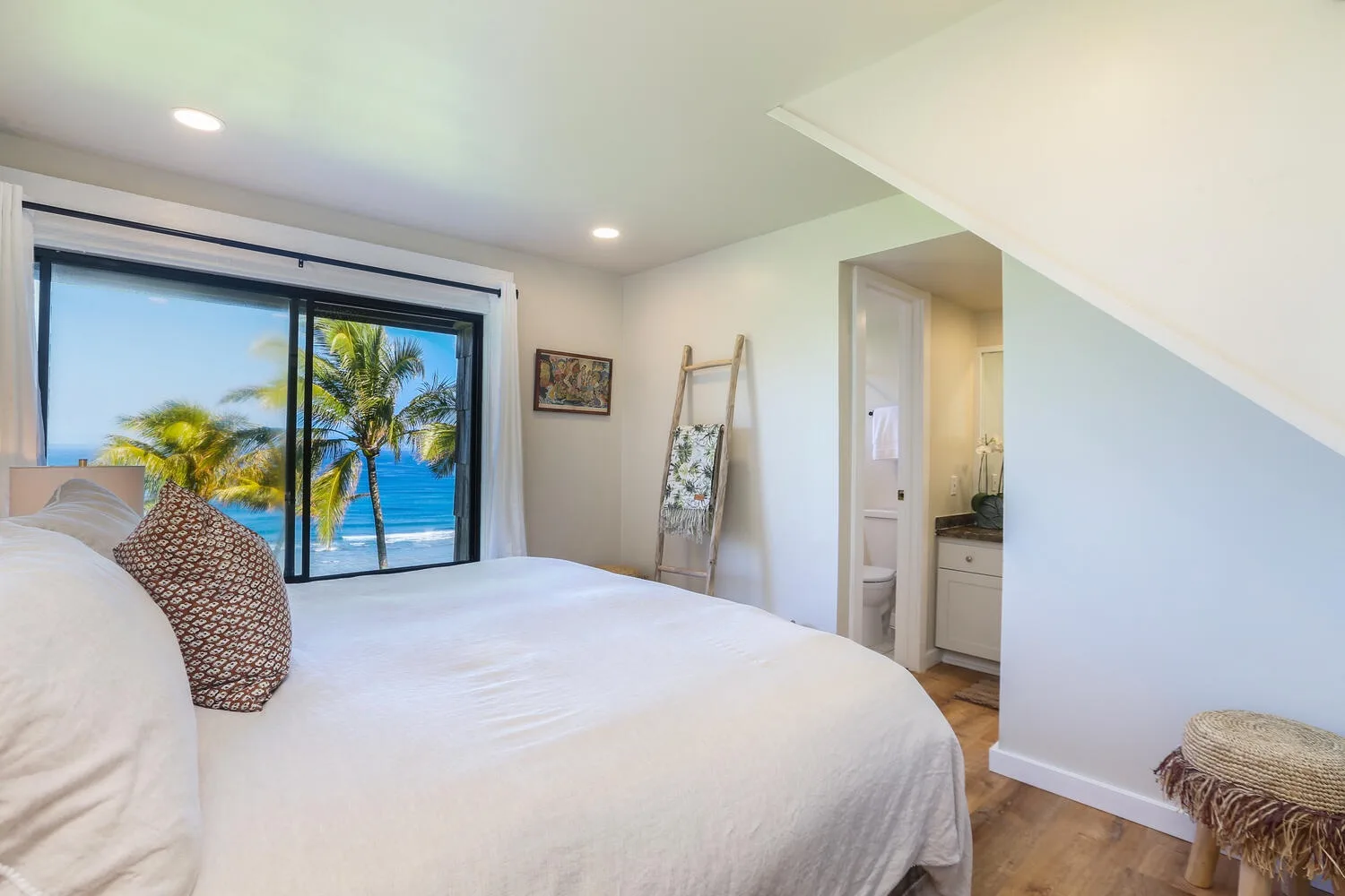 Un dormitorio con una cama blanca junto a una ventana amplia con vista a palmeras y al mar.
