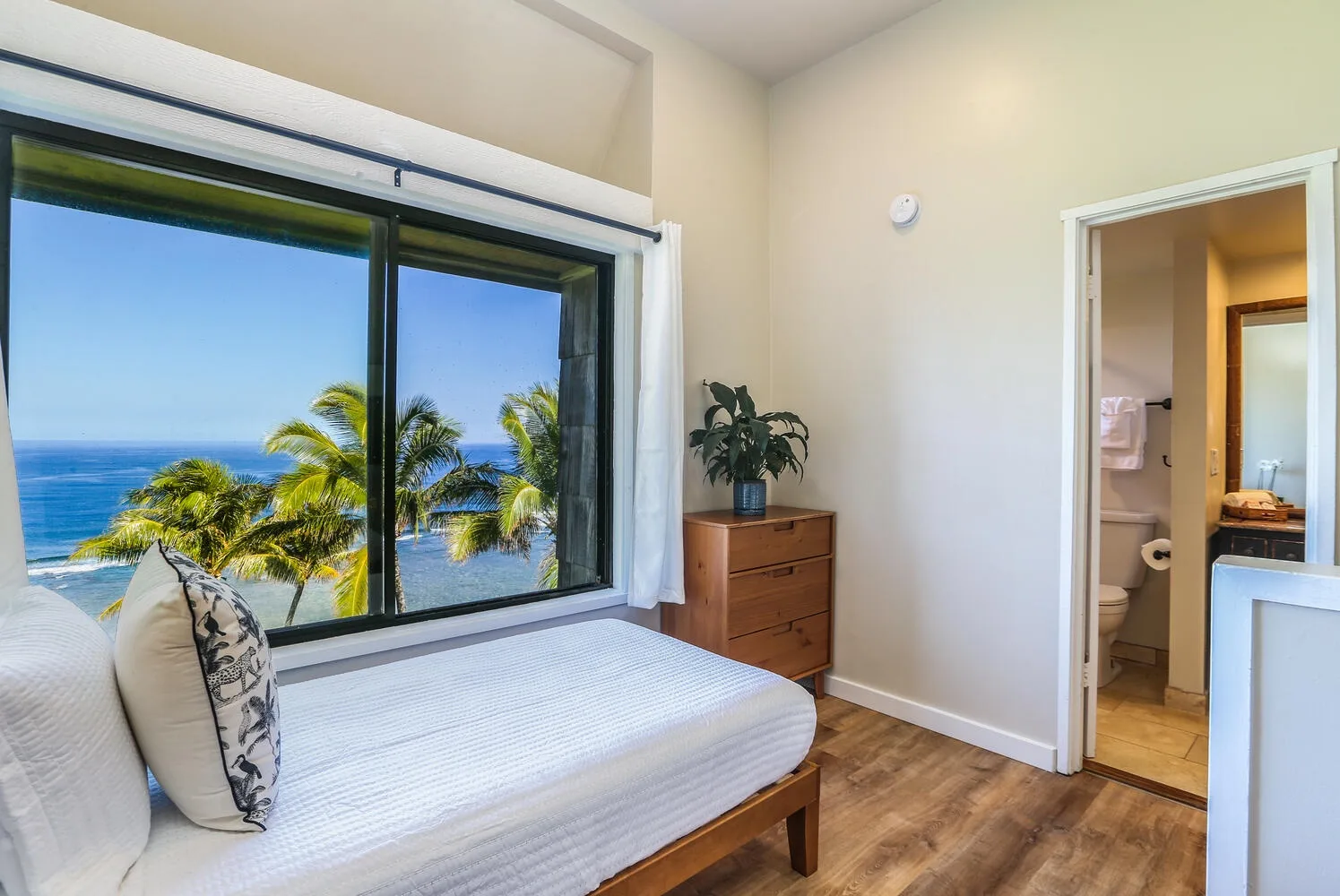 Un dormitorio con una cama junto a una ventana grande con vista a palmeras y al océano.
