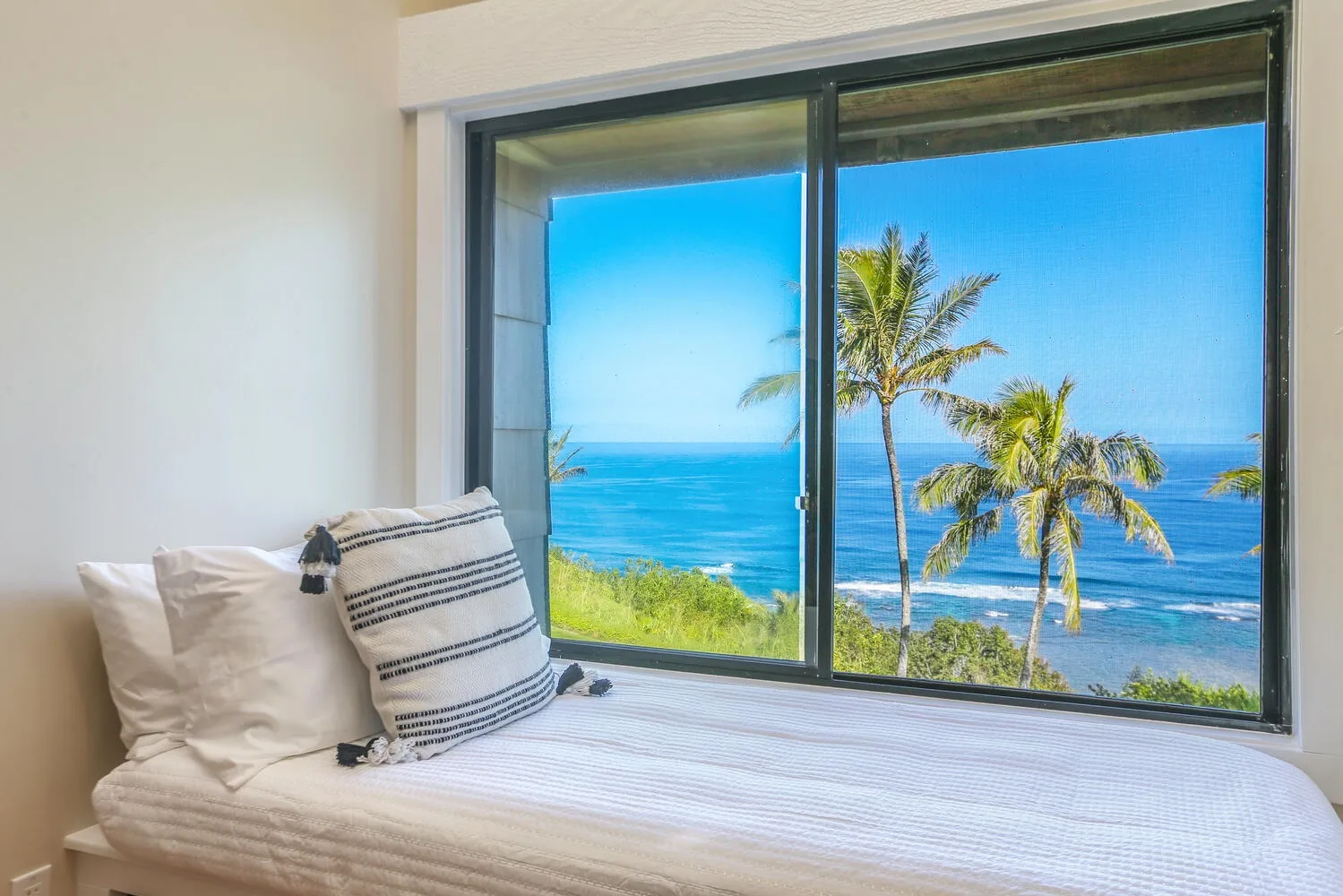 Un dormitorio con una cama junto a una ventana grande con vistas al océano y palmeras.
