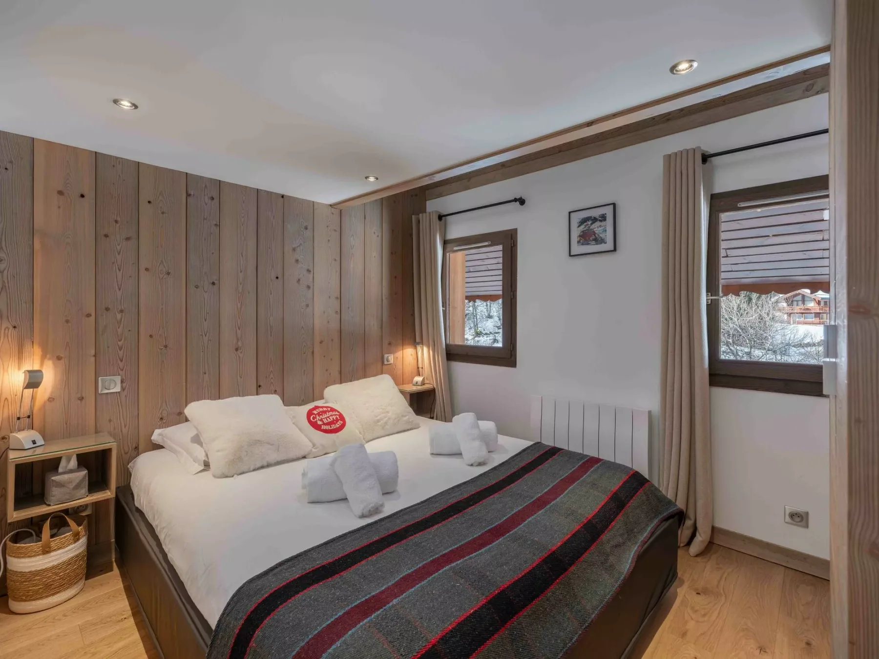 A bedroom with a double bed, wooden wall paneling, and two windows.