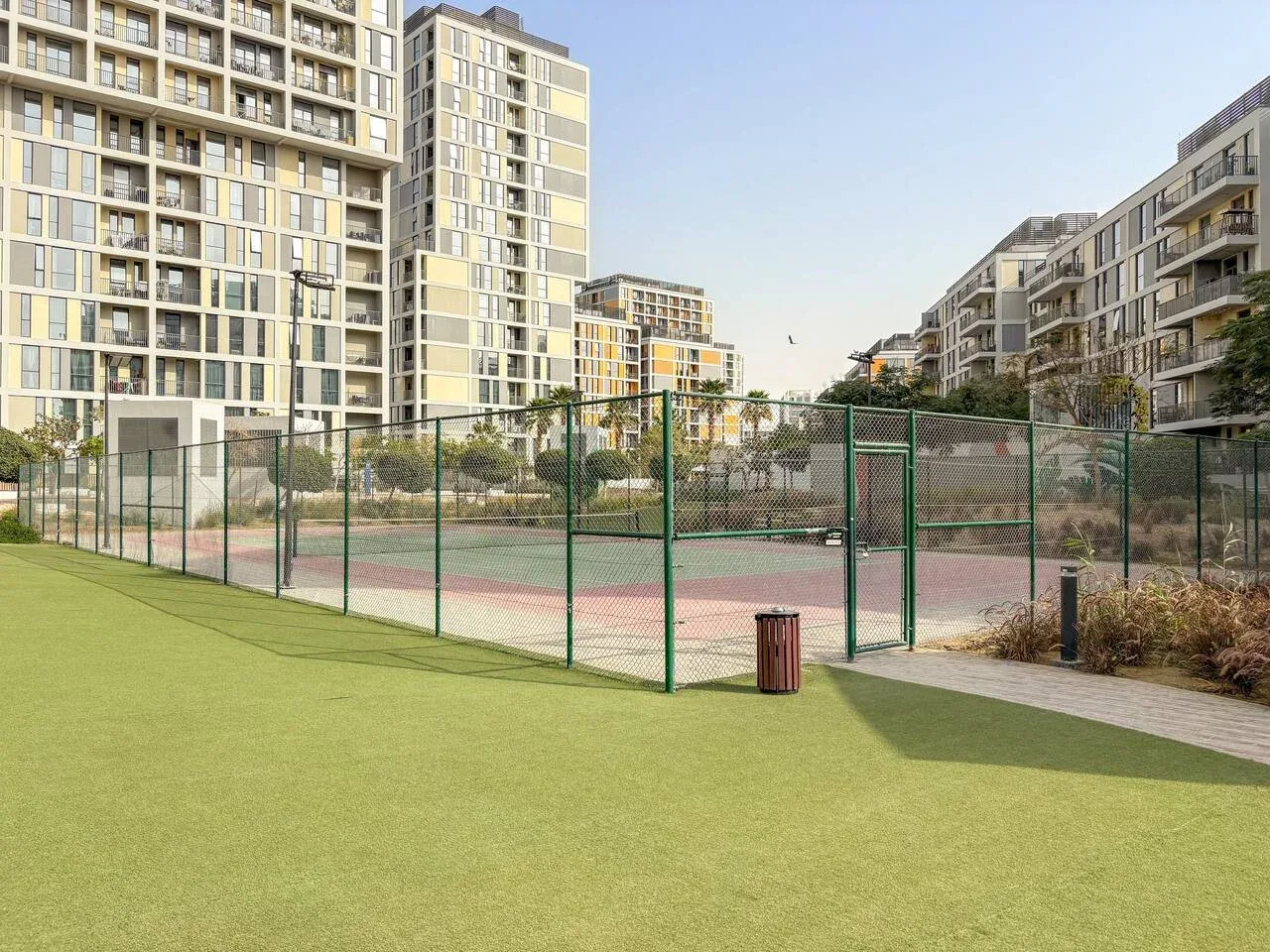 An outdoor sports court with green fencing sits between modern apartment buildings.