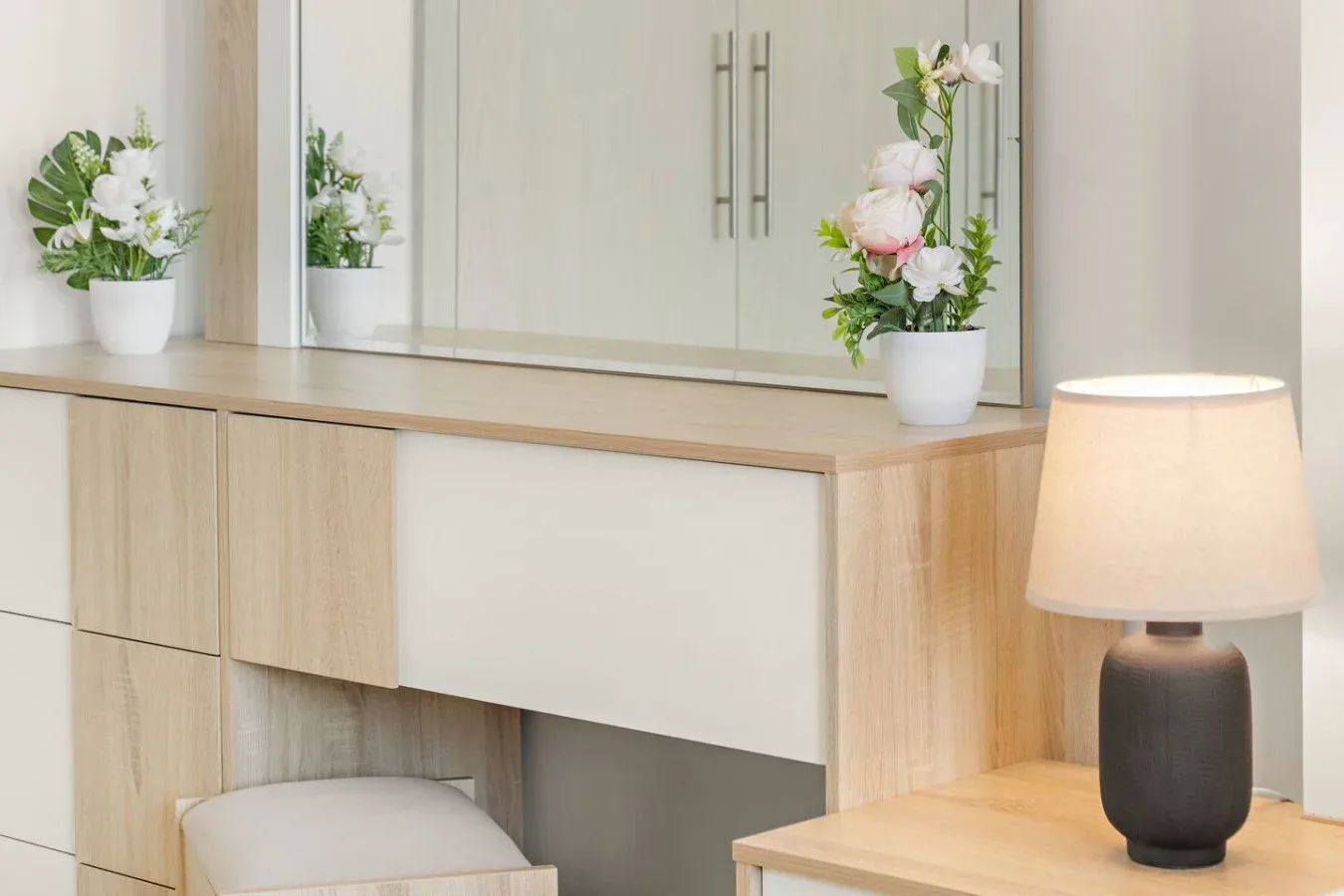 A bedroom vanity with a mirror, flower arrangements, and a lit table lamp.