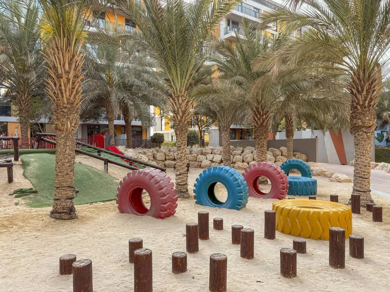 A landscaped outdoor play area has colorful tire-shaped play structures among palm trees.