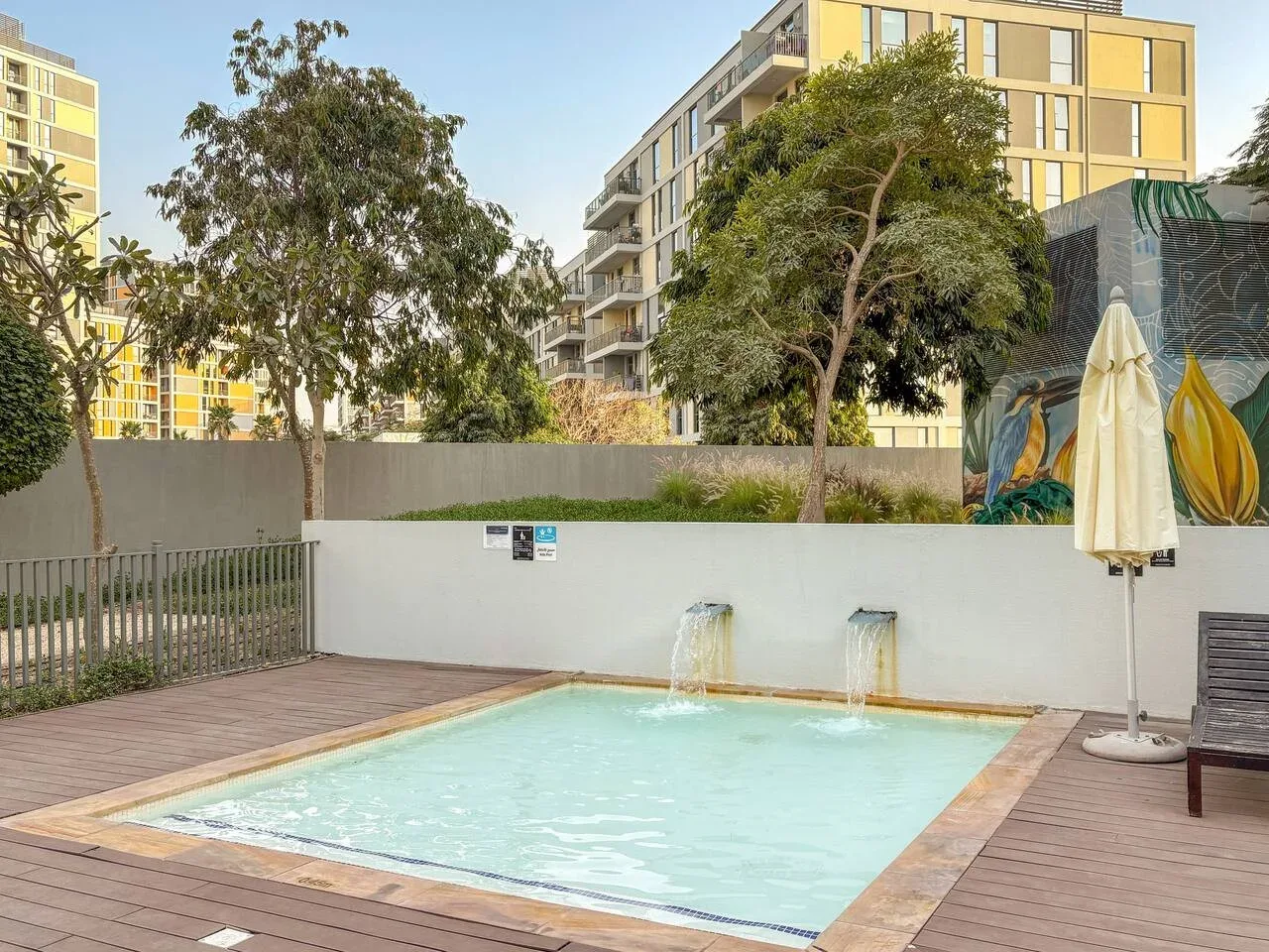 An outdoor swimming pool sits on a wooden deck beside a low wall and trees.