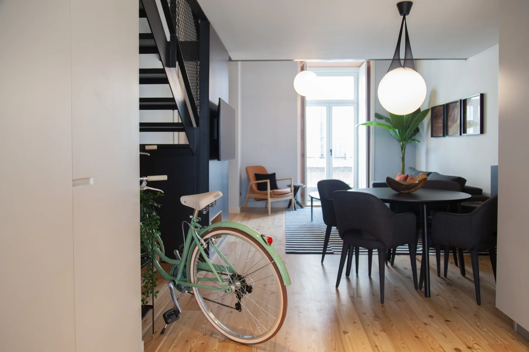 Zona de comedor y estar con asientos oscuros, una lámpara colgante y una bicicleta junto a la escalera.