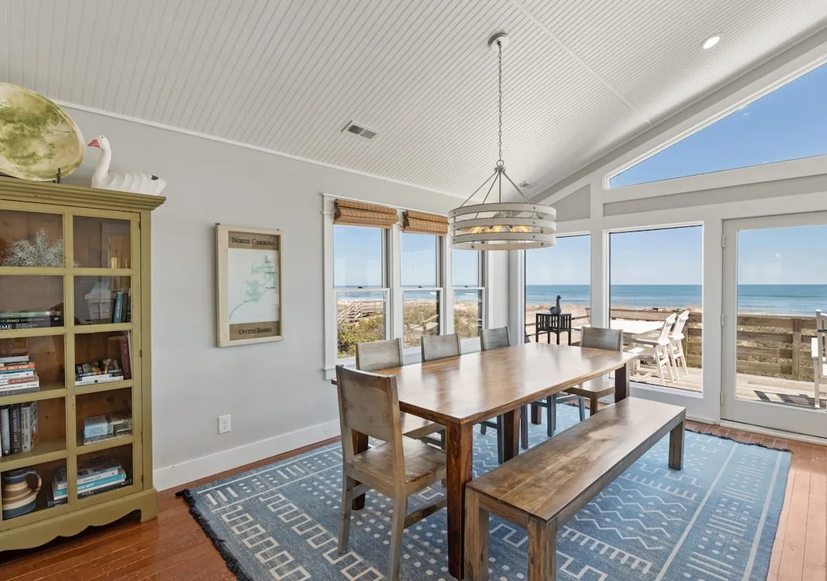 Un comedor luminoso con una mesa de madera y grandes ventanas con vistas al océano.
