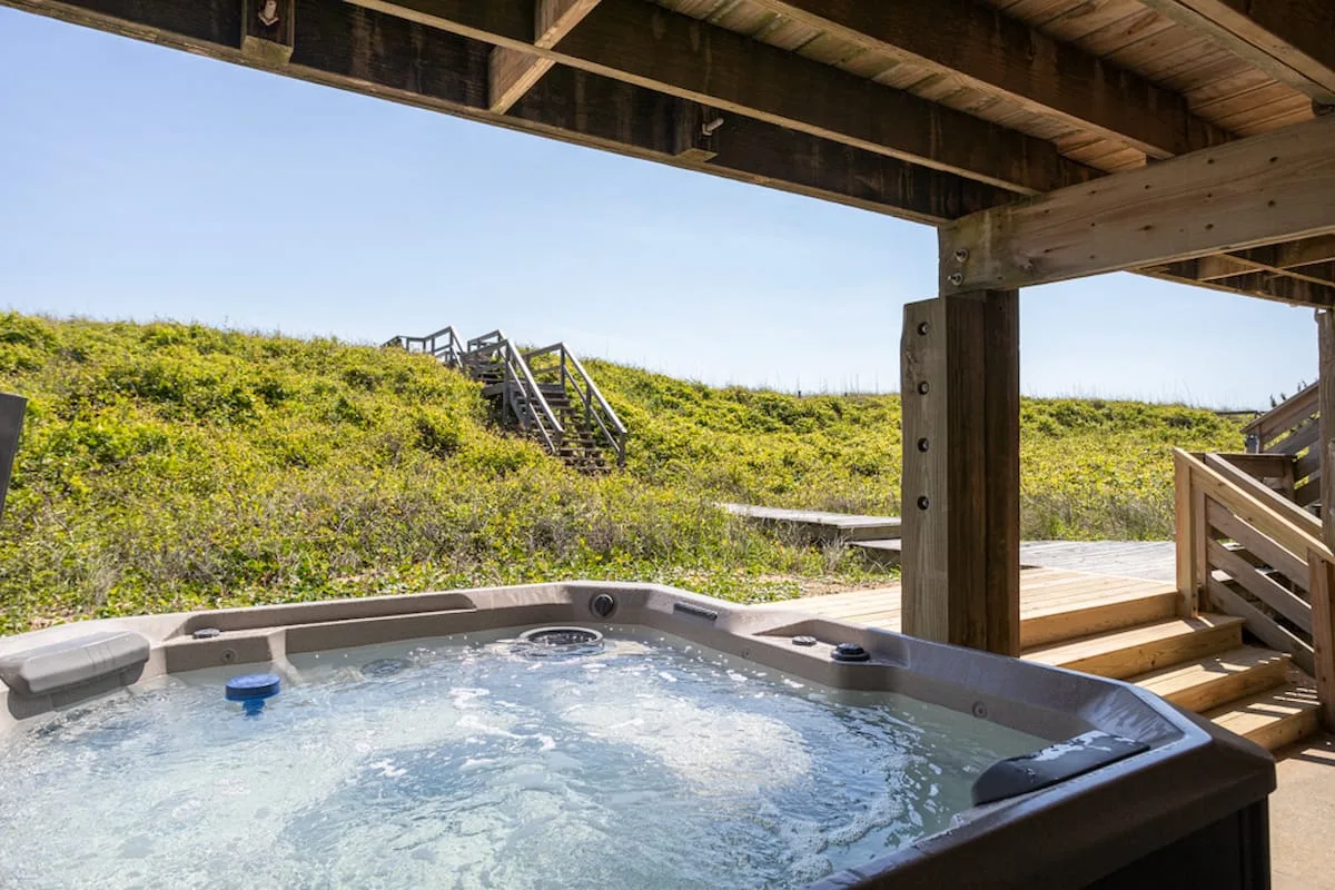 Una bañera de hidromasaje al aire libre está bajo una terraza de madera junto a dunas cubiertas de hierba.