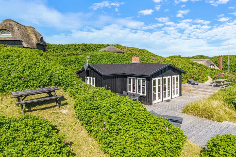 Una casa oscura se encuentra entre dunas verdes, junto a una terraza de madera y una mesa de picnic.