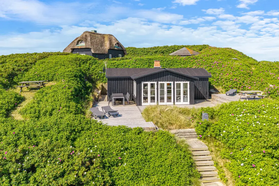 Una casa oscura con terraza y escalones se encuentra entre dunas verdes.