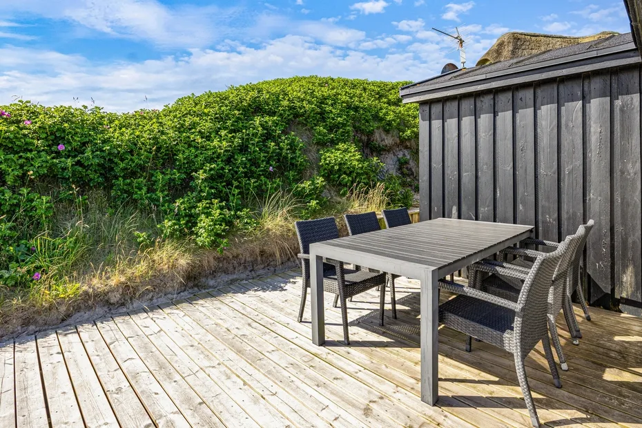 Zona de comedor al aire libre con mesa y asientos en una terraza de madera junto a un jardín.