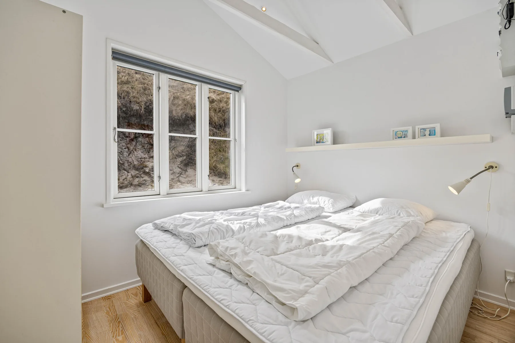 Un dormitorio luminoso con una cama bajo una ventana y un estante estrecho en la pared.
