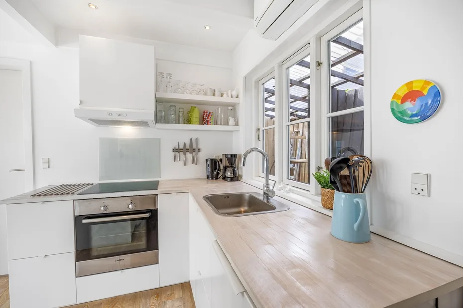 Una cocina blanca y luminosa con fregadero, placa de cocina y ventanas grandes.