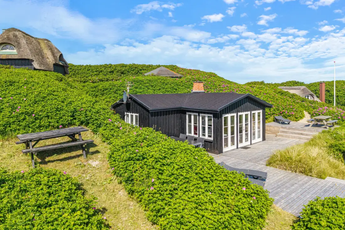 Una casa oscura se encuentra entre la vegetación de las dunas, junto a una terraza de madera.