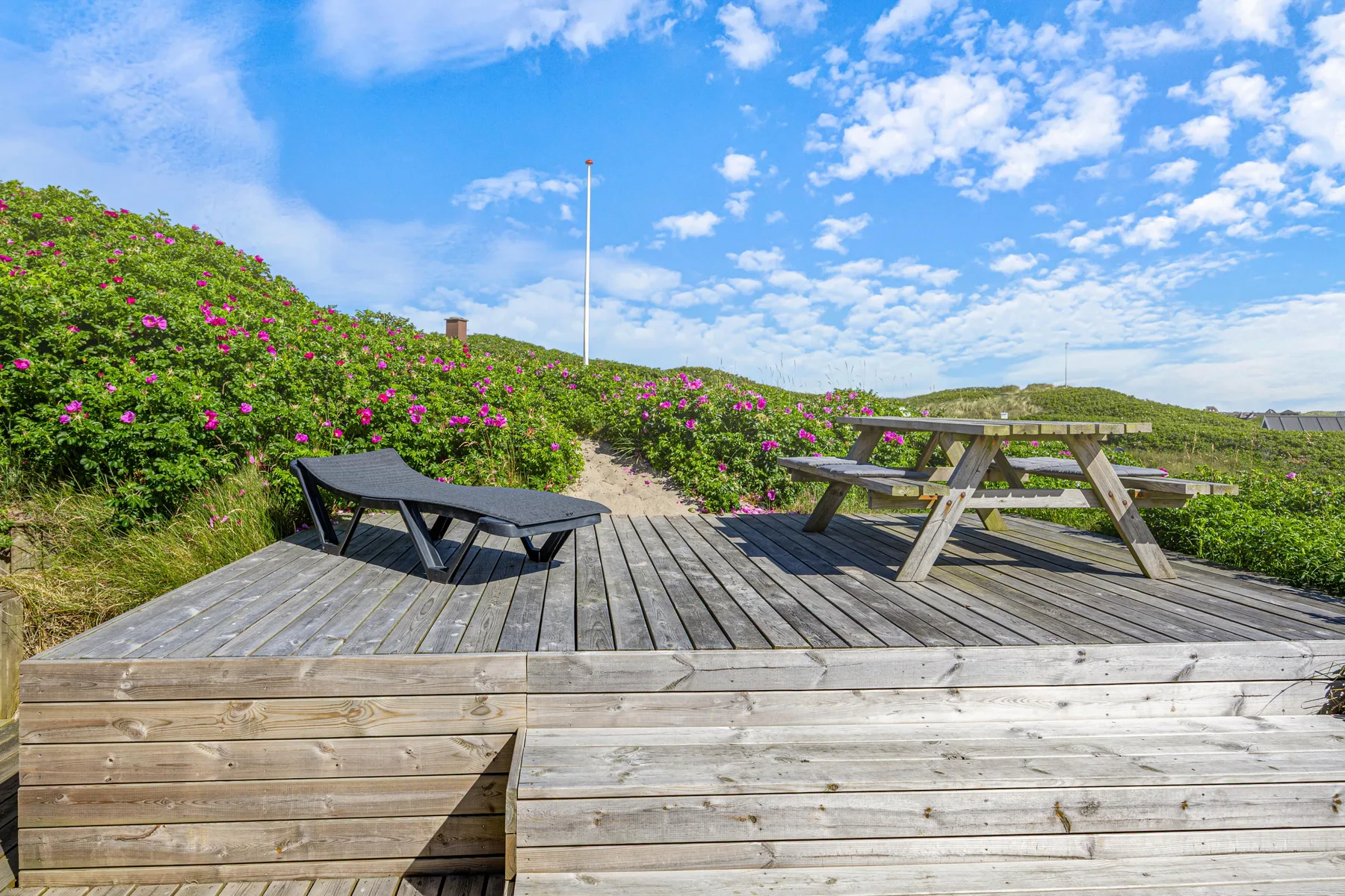 Una terraza elevada de madera tiene asientos y una mesa de picnic entre arbustos en flor.