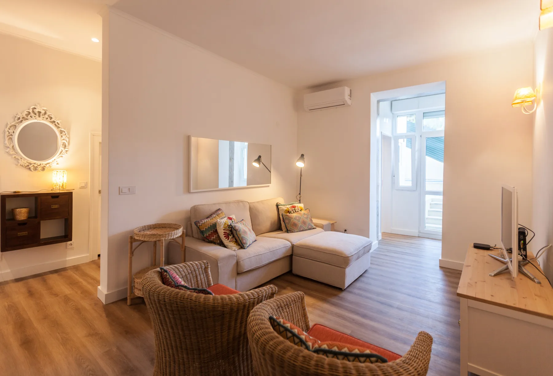 A bright living room with seating, a wall mirror, and a glass door to a balcony.