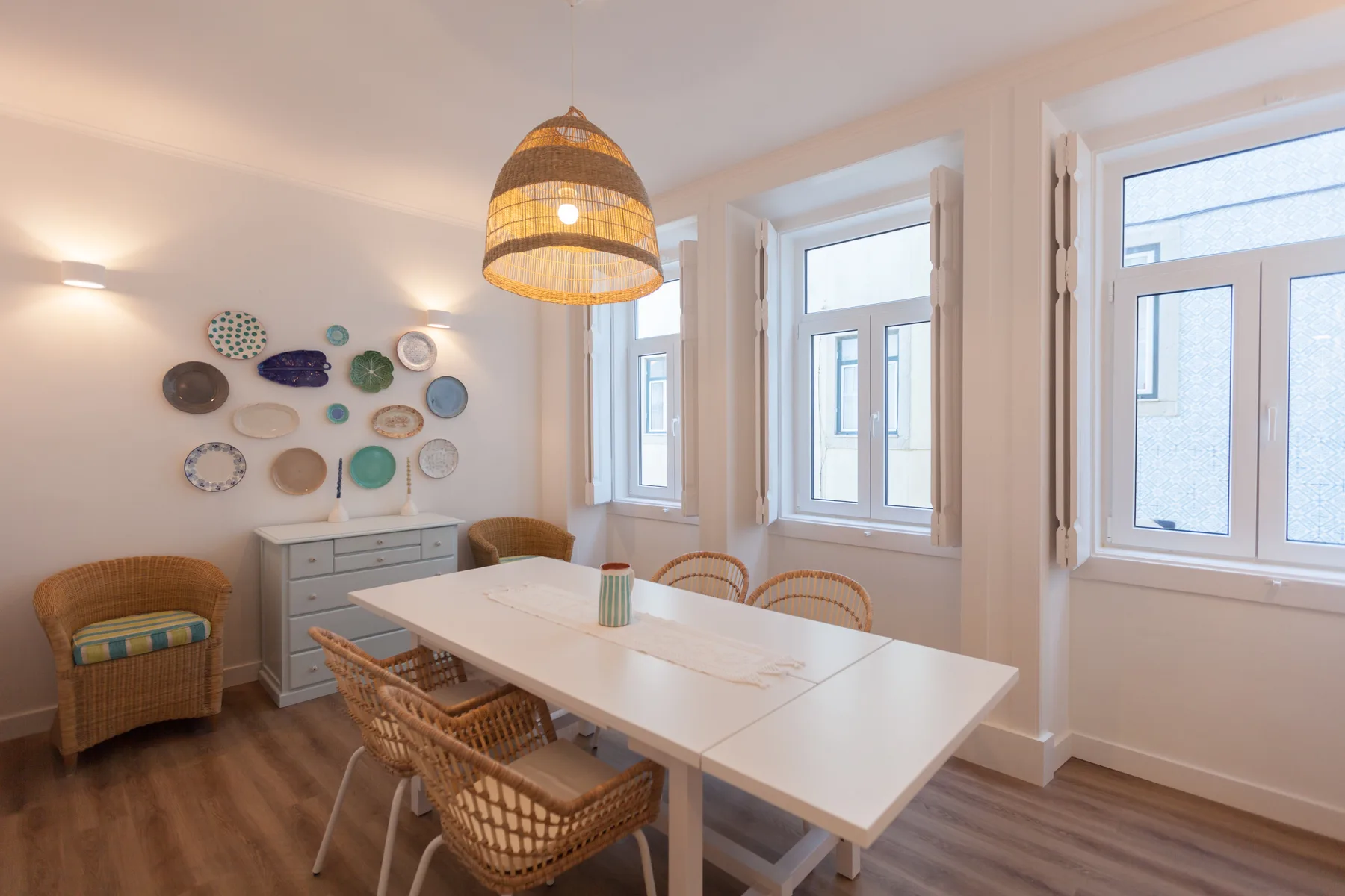 A dining area with a white table, woven seating, and several windows.