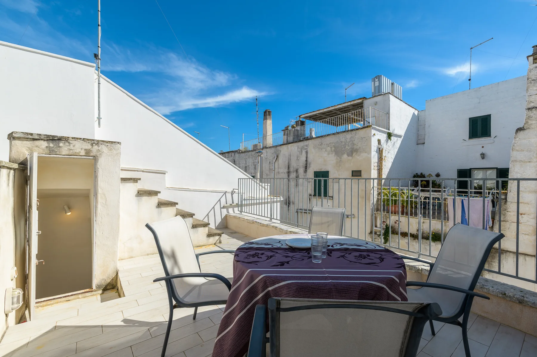 A rooftop terrace has a round table and several chairs beside a metal railing.