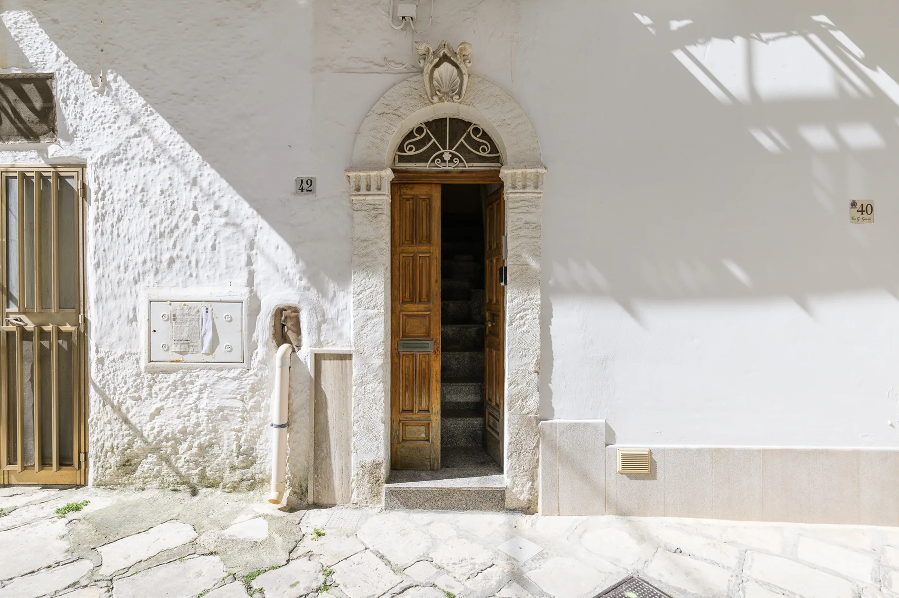 A sunlit white building facade with an ornate arched doorway and open wooden door.