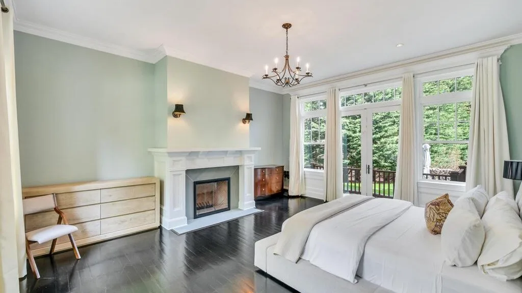 Un dormitorio luminoso con una cama blanca junto a una chimenea y ventanas altas con vistas al jardín.