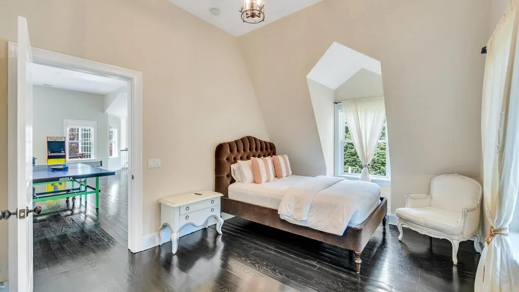 Un dormitorio con una cama, una mesita de noche y un sillón junto a una ventana alta.