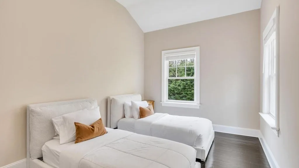 Un dormitorio luminoso con dos camas, paredes neutras y ventanas con vistas a la vegetación.