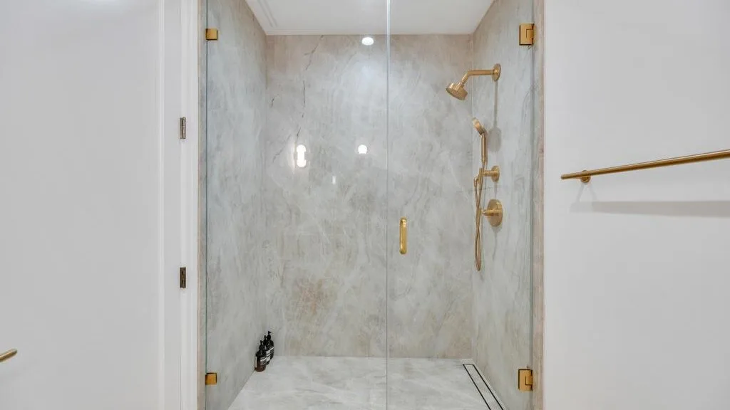 Una ducha con azulejos de mármol, una mampara de vidrio y accesorios dorados.