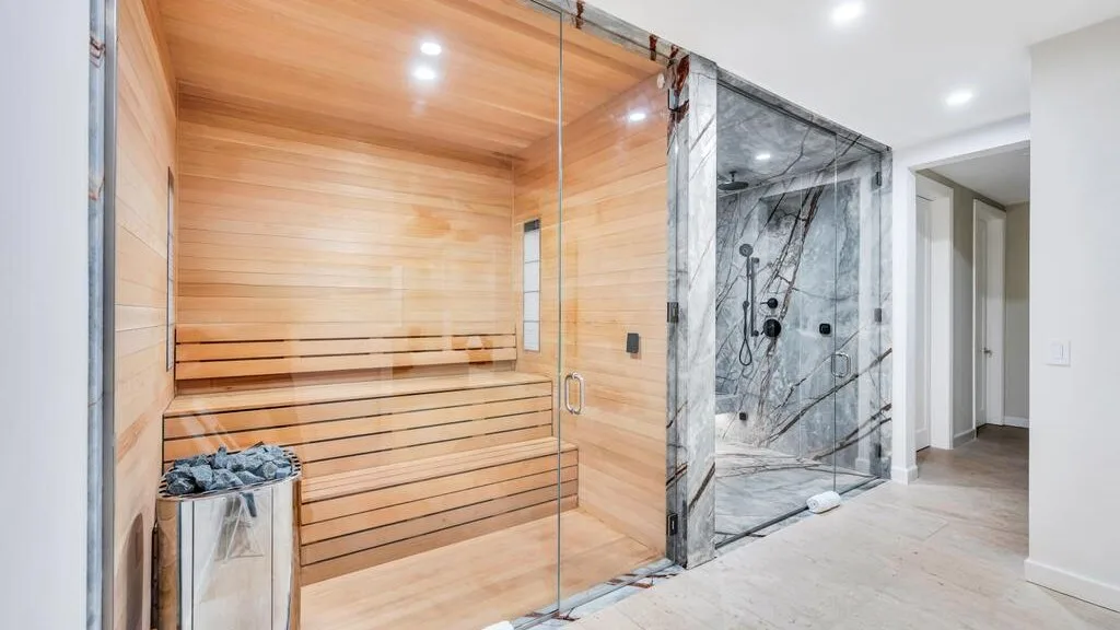 Una sauna revestida de madera está junto a una ducha con mampara de vidrio en un baño luminoso.