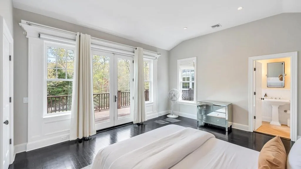 Un dormitorio luminoso con una cama junto a ventanas altas y un mueble con frente de vidrio.