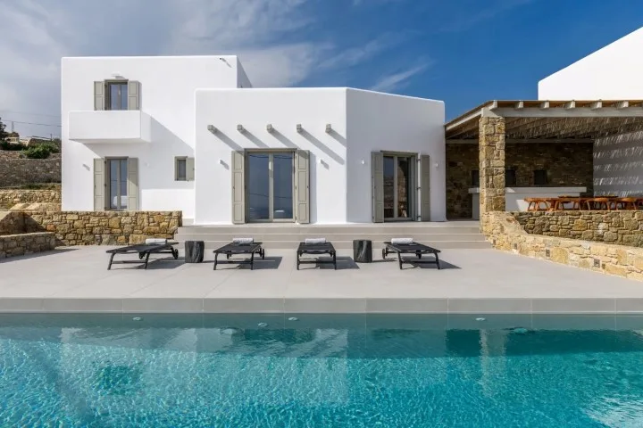 A modern white house stands beside a turquoise swimming pool and outdoor seating.