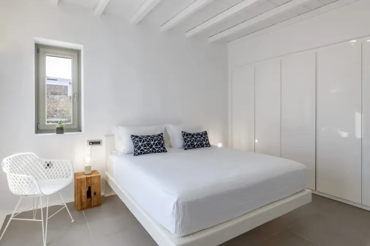 A bright bedroom with a white bed beside a window and built-in wardrobes.