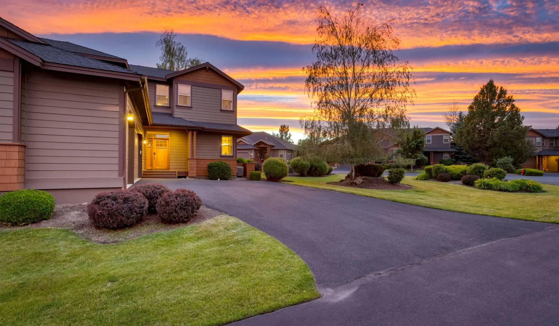 A paved road curves past residential buildings and landscaped lawns beneath a colorful sunset sky.
