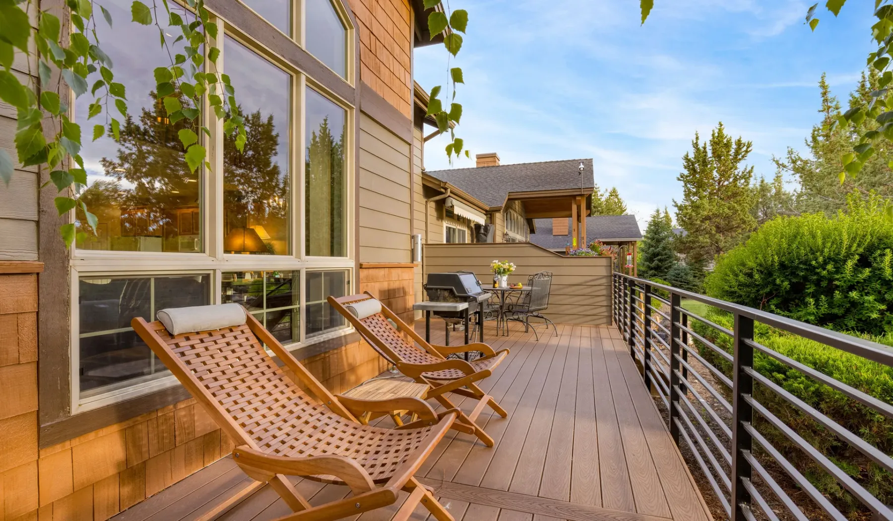 A wooden deck with reclining seating overlooks leafy trees beside large windows.