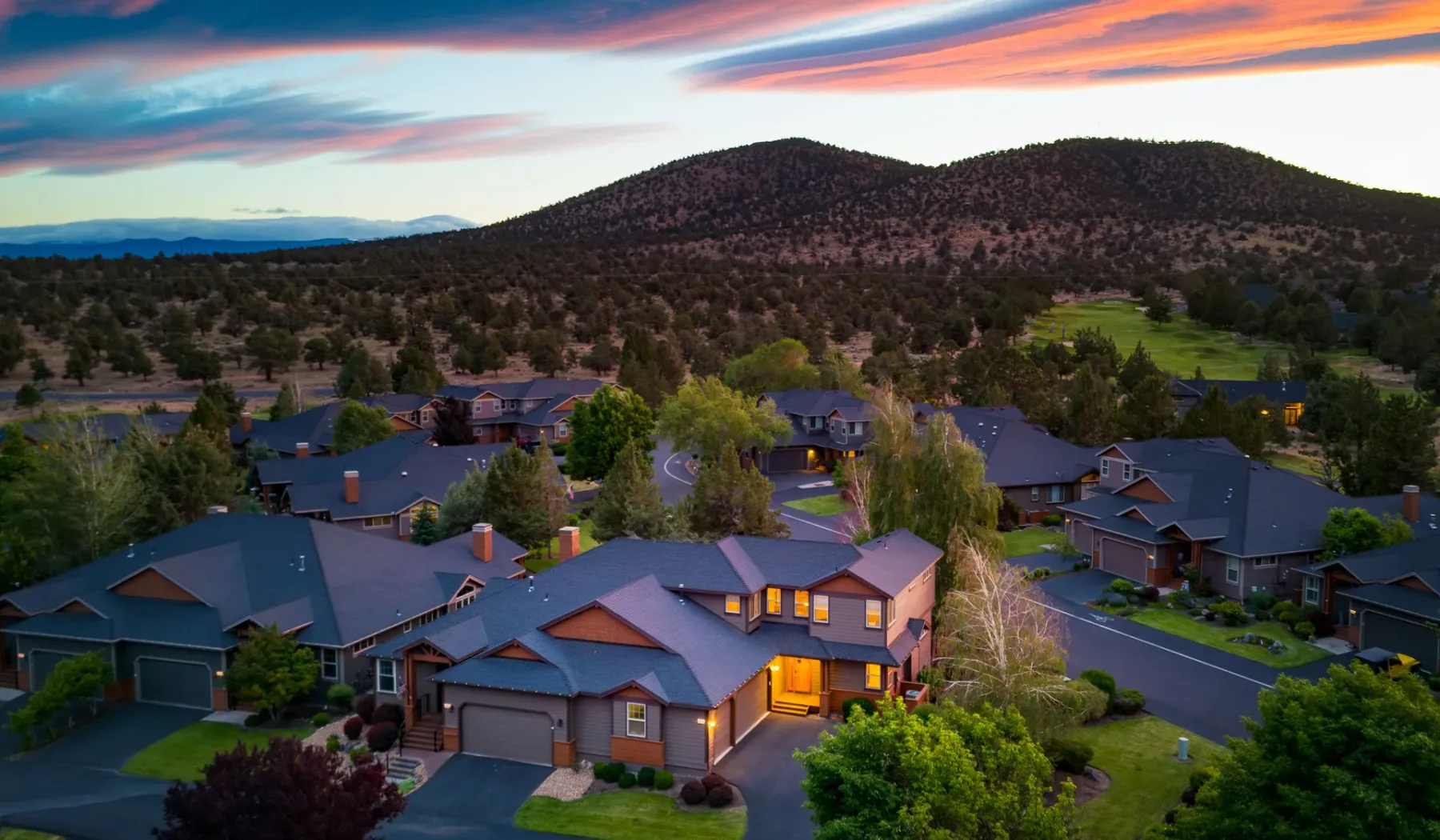 A residential neighborhood sits among trees beneath a low mountain ridge at sunset.
