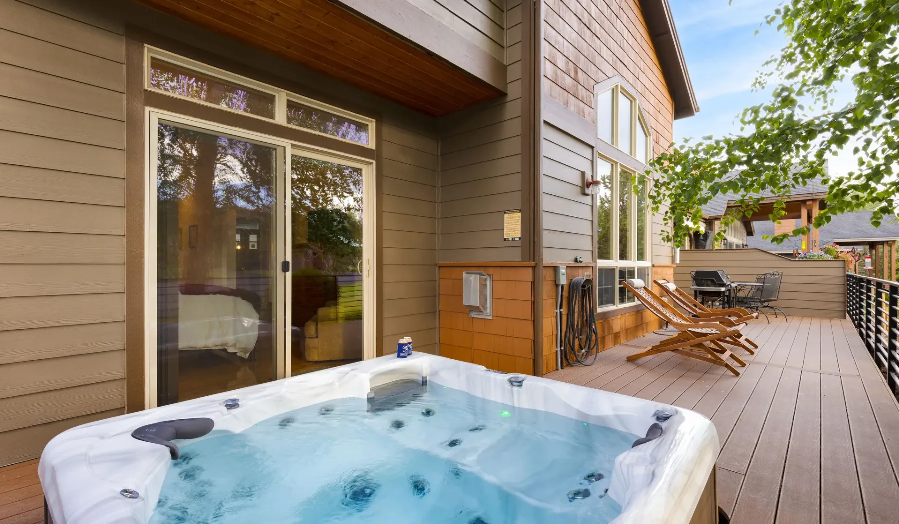 An outdoor hot tub sits on a wooden deck beside a house and patio seating.