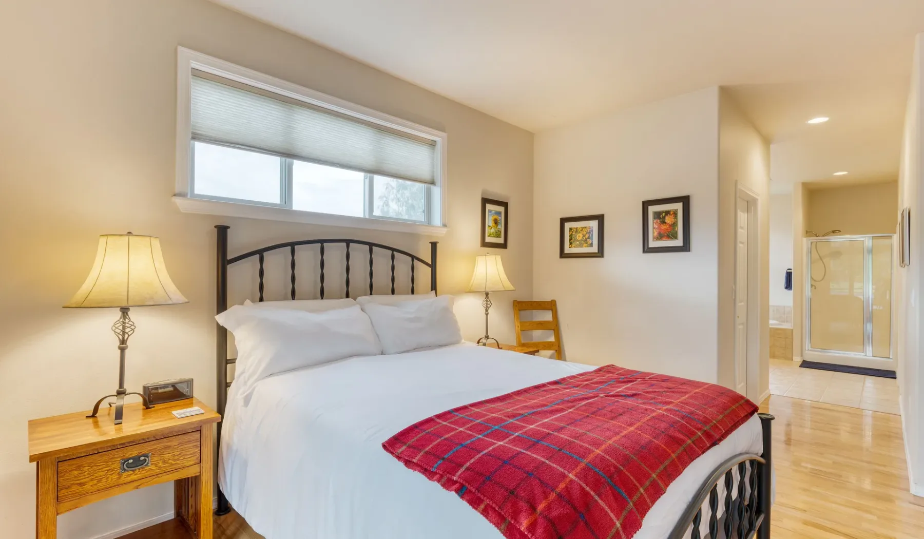 A bedroom with a white bed, red plaid blanket, and windowside seating.