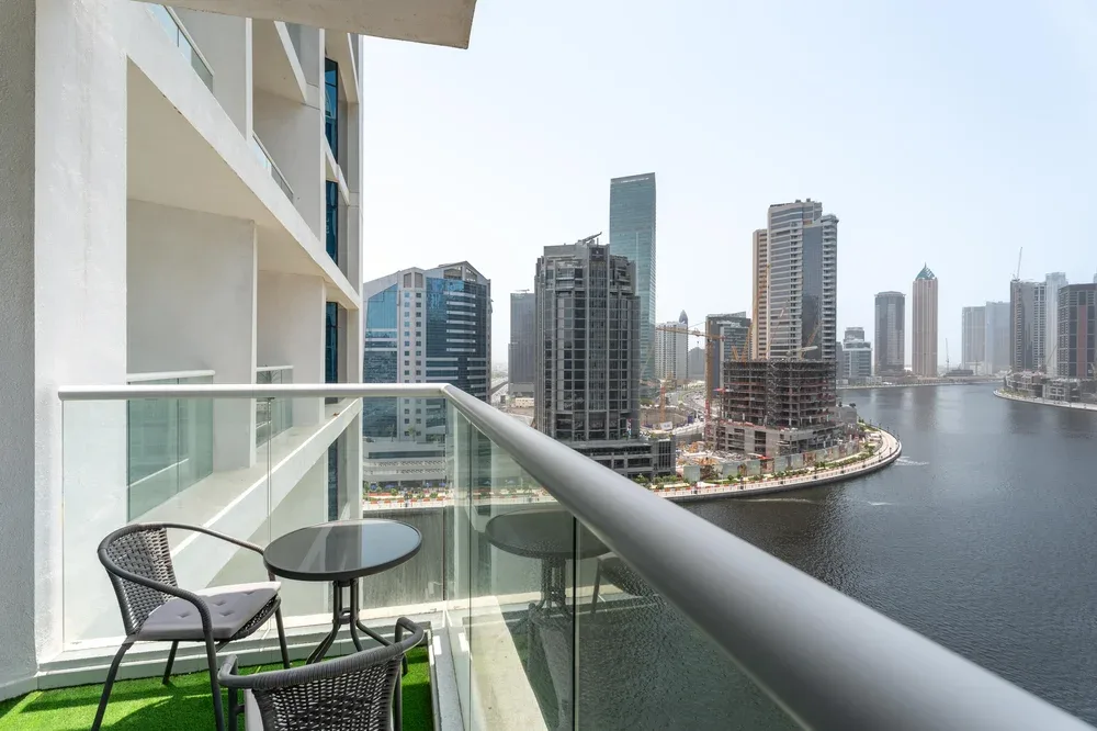 A balcony overlooks a waterfront skyline, with a small seating area beside the glass railing.