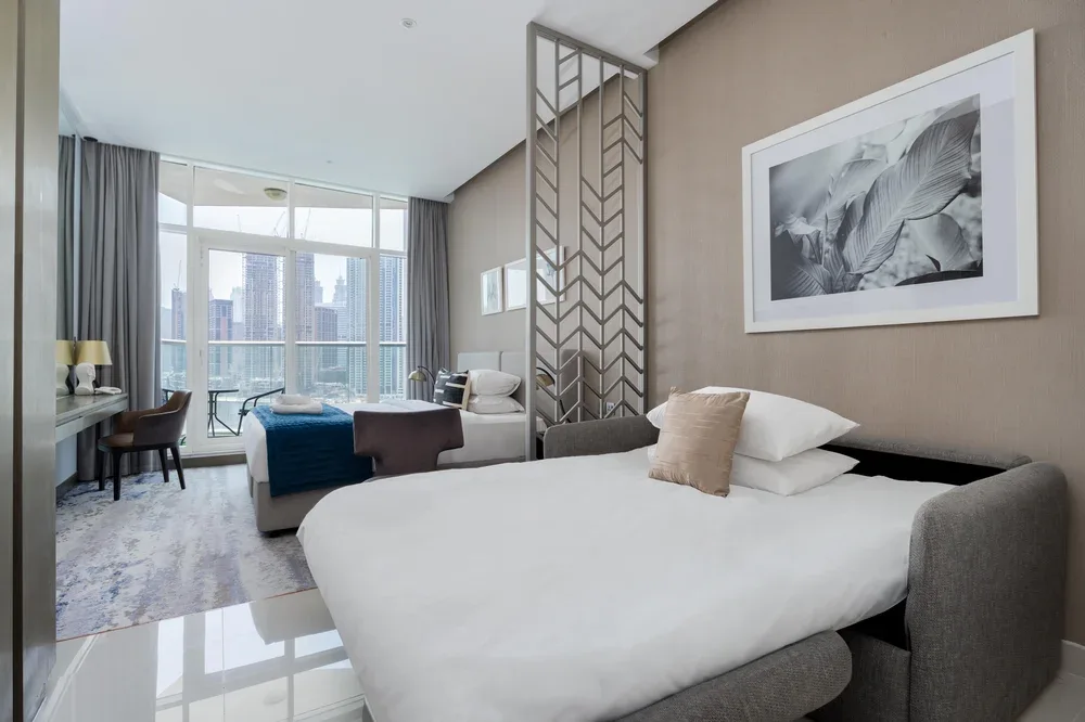 A bedroom with a large bed, seating area, and floor-to-ceiling windows overlooking city buildings.