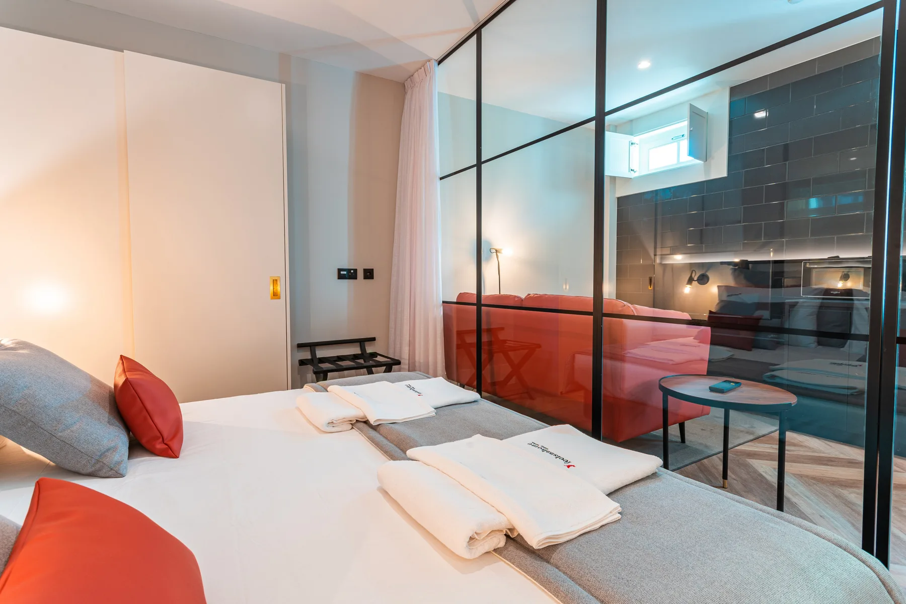 A bedroom with white bedding and a glass partition overlooking the kitchen.